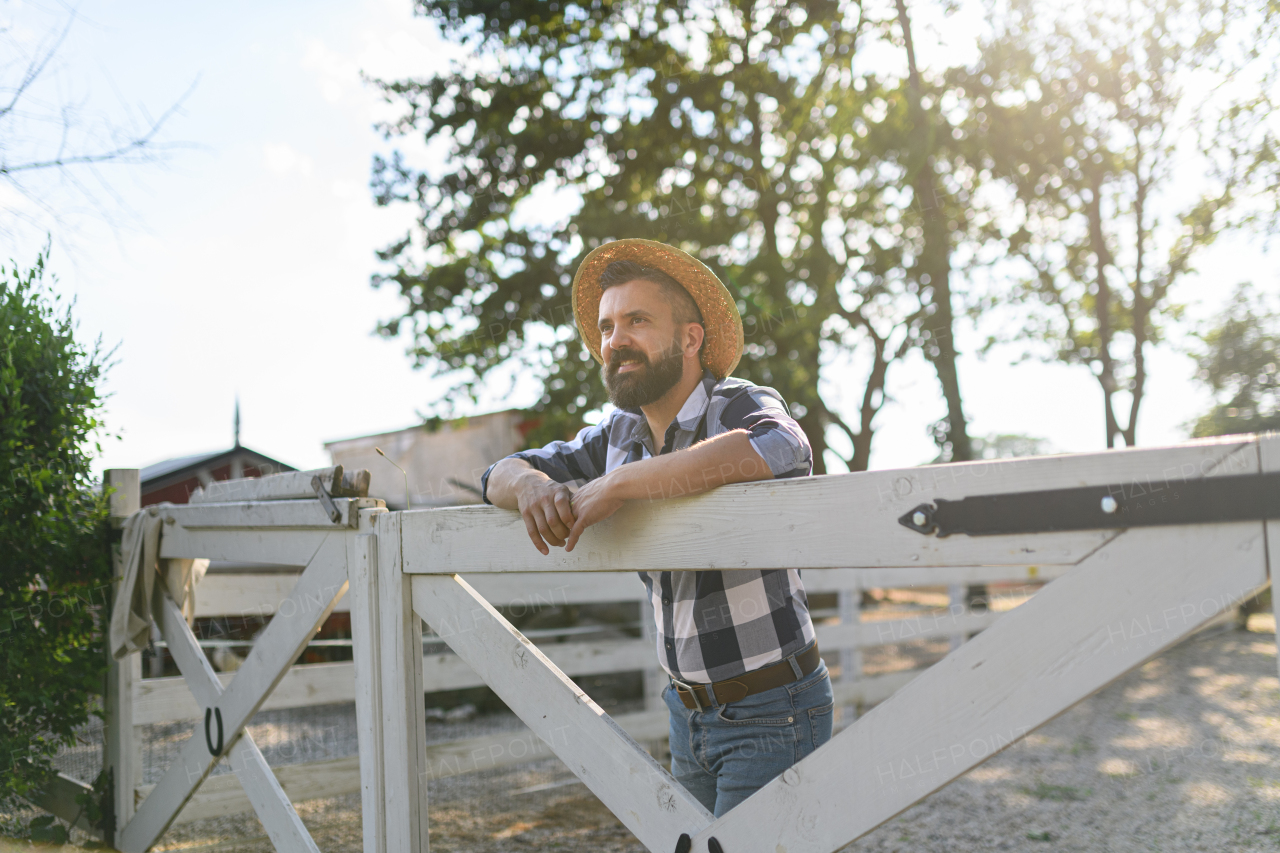 Portrait of handsome farmer leaning agaist wooden fence, looking at his farm. Man with hat working on family ranch.