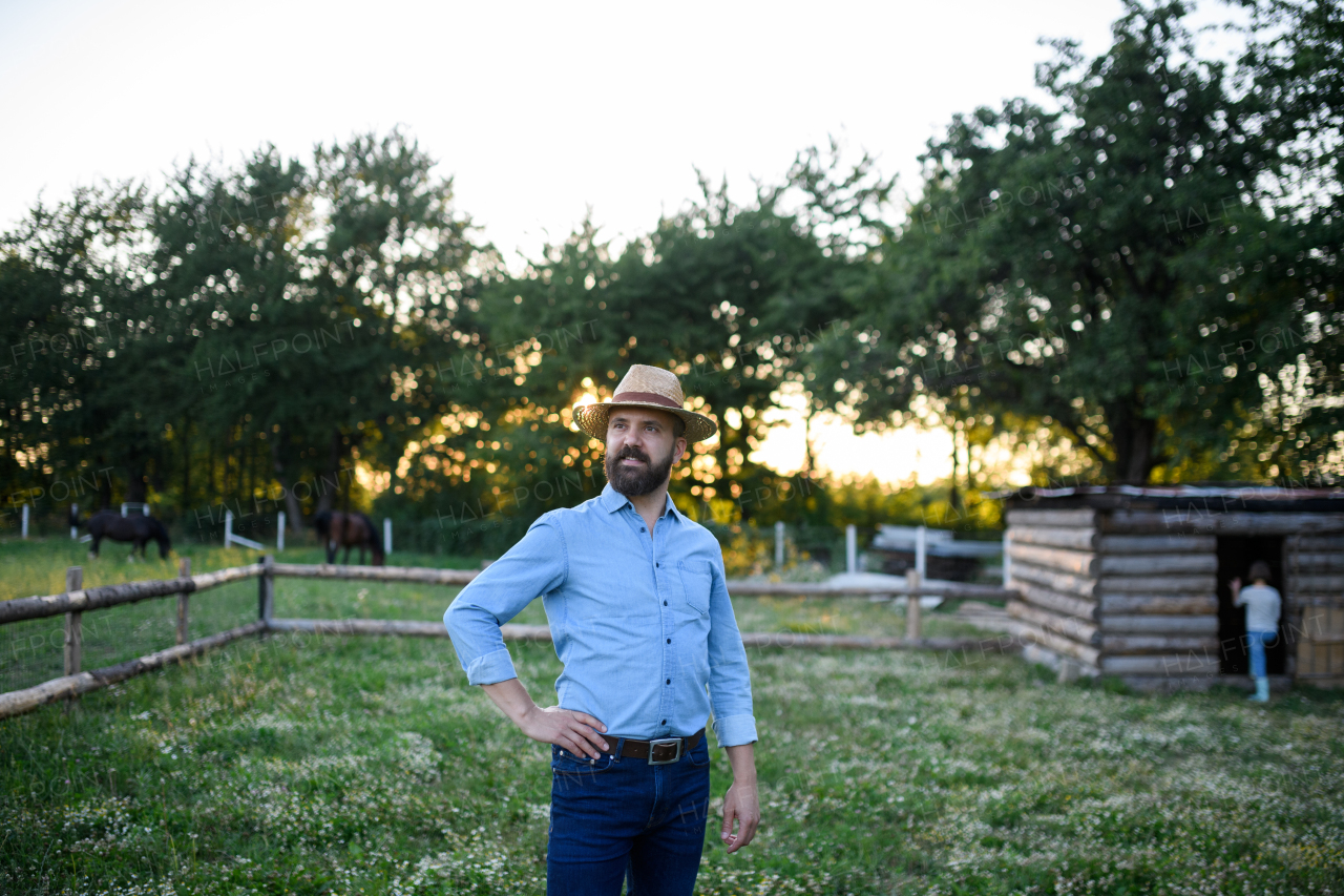 A portrait of happy mature man farmer with hat standing outdoors on family farm at sunset.