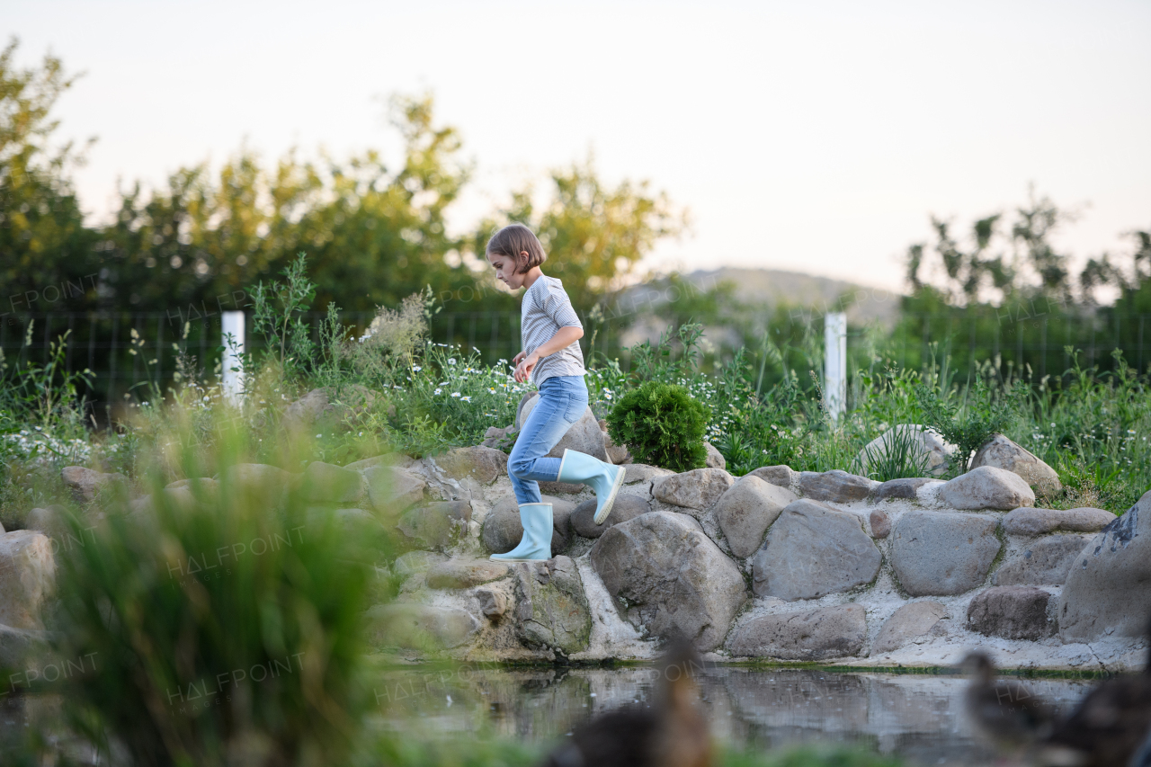 Beautiful young girl in rubber boots running by pond, helping on family farm during summer.