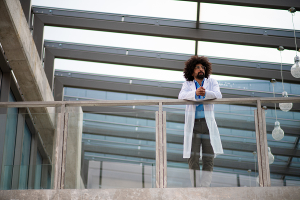 Portrait of male doctor in white coat standing in hospital, leaning on glass railing, looking at camera.