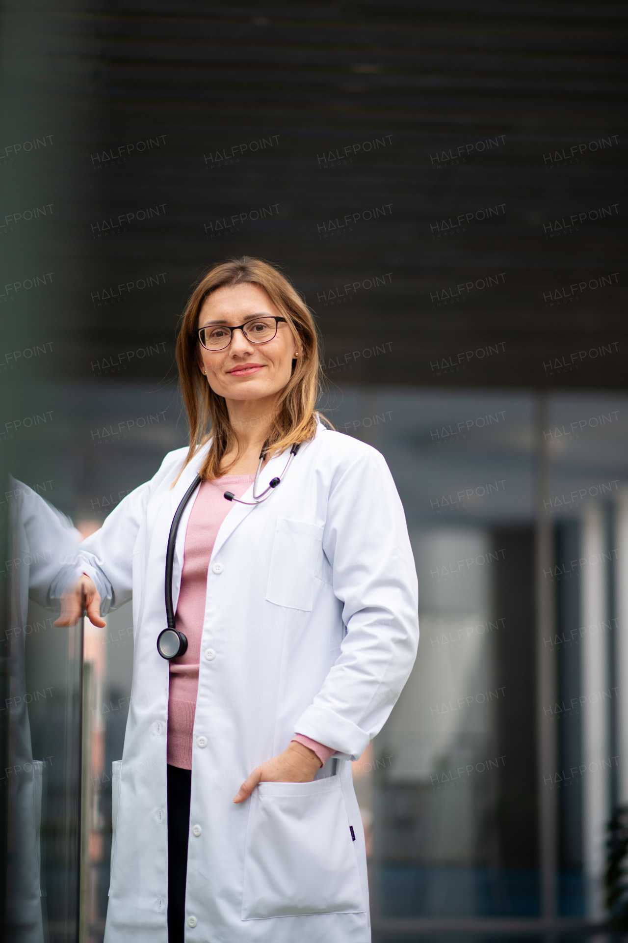 Portrait of female doctor in white coat standing in hospital, leaning on glass railing, looking at camera.