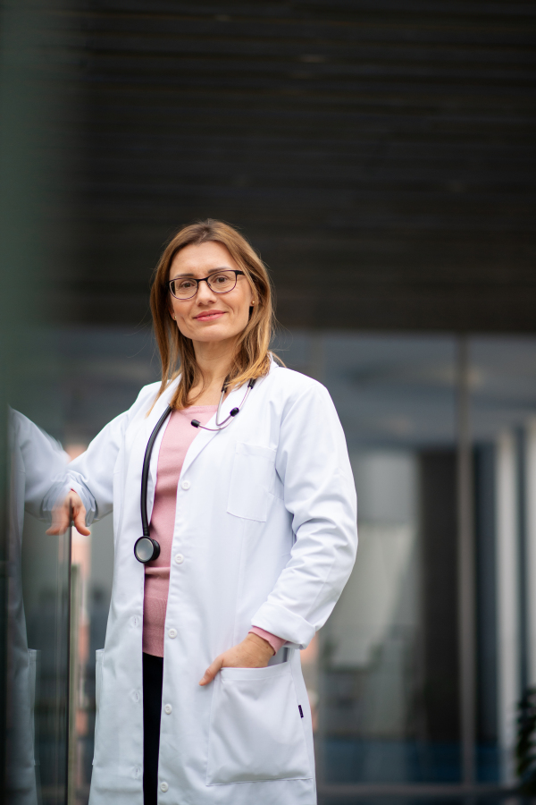 Portrait of female doctor in white coat standing in hospital, leaning on glass railing, looking at camera.