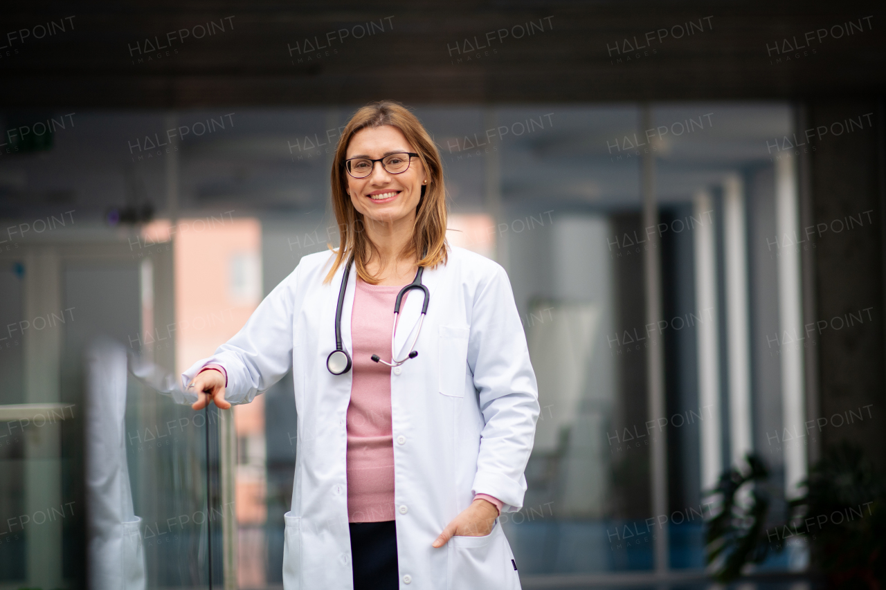 Portrait of female doctor in white coat standing in hospital, leaning on glass railing, looking at camera.