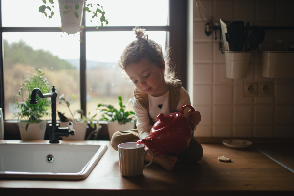 Adorable little girl is pouring tea from the teapot into cup. The sick girl with a cold is sitting on the kitchen counter and concentrating.