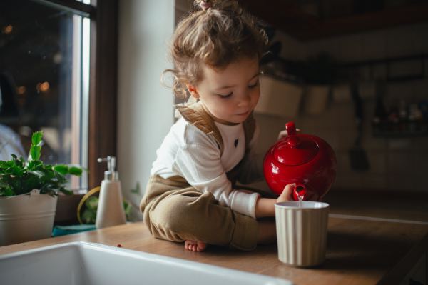 Adorable little girl is pouring tea from the teapot into cup. The sick girl with a cold is sitting on the kitchen counter and concentrating.