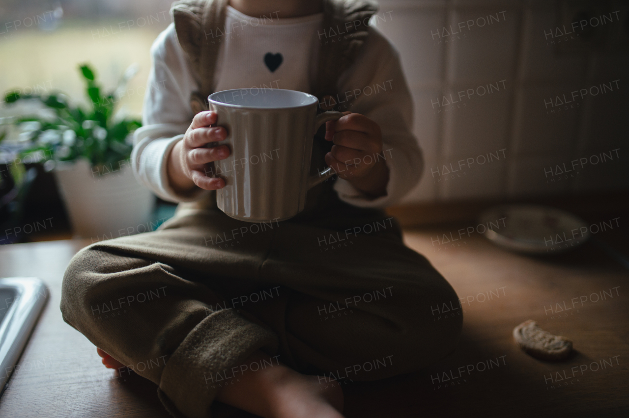 Adorable little girl is drinking tea. The sick girl with cold is sitting on kitchen counter with mug in hand