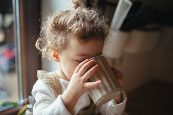 Adorable little girl is pouring tea from the teapot into cup. The sick girl with a cold is sitting on the kitchen counter and concentrating.