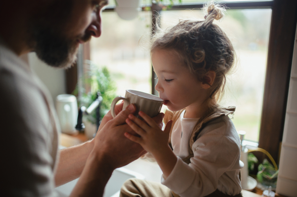 Father helping little daughter to drink warm tea from cup. Sick girl with cold is sitting on kitchen counter and drinking herbal tea