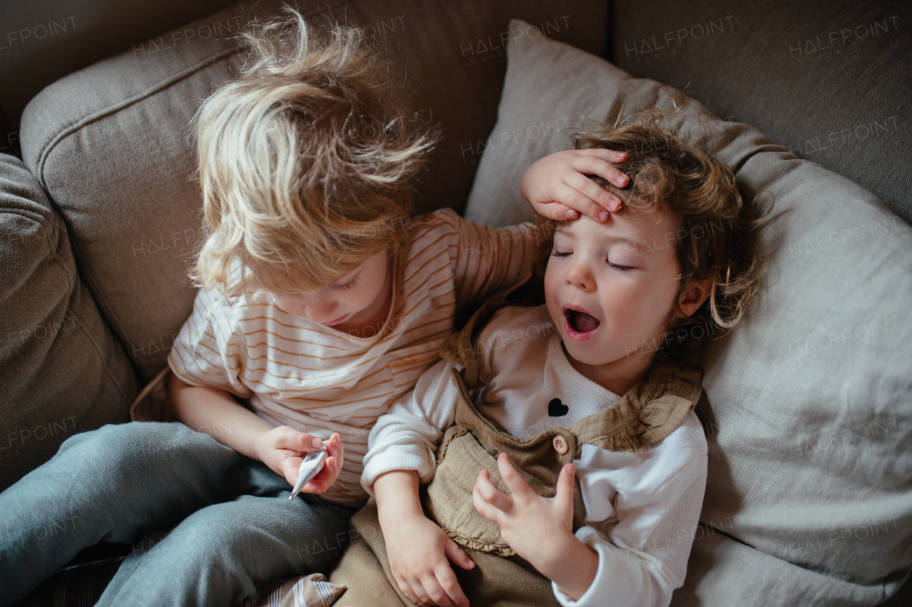 Siblings sitting on sofa, brother touching sick sister's forehead, checking her temperature