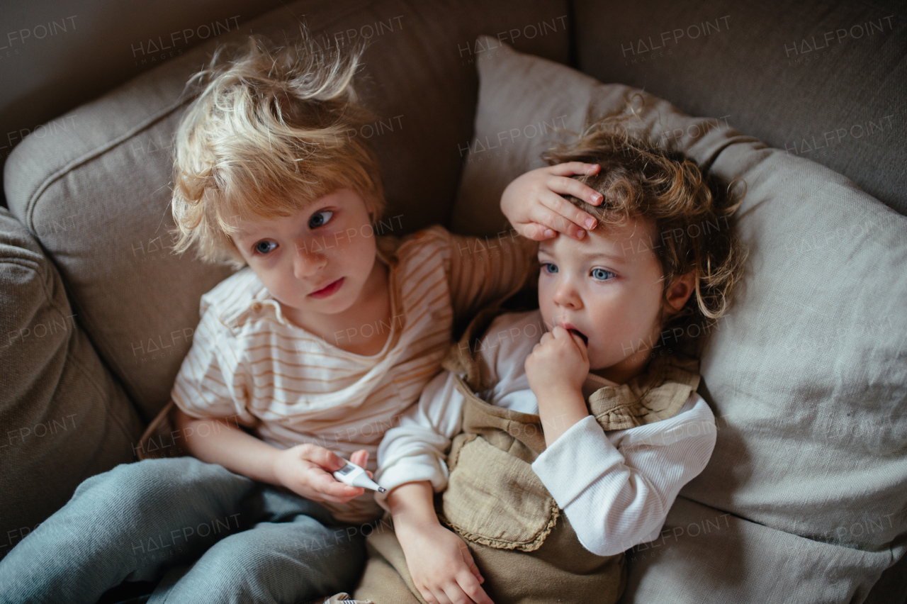 Siblings sitting on sofa, brother touching sick sister's forehead, checking her temperature