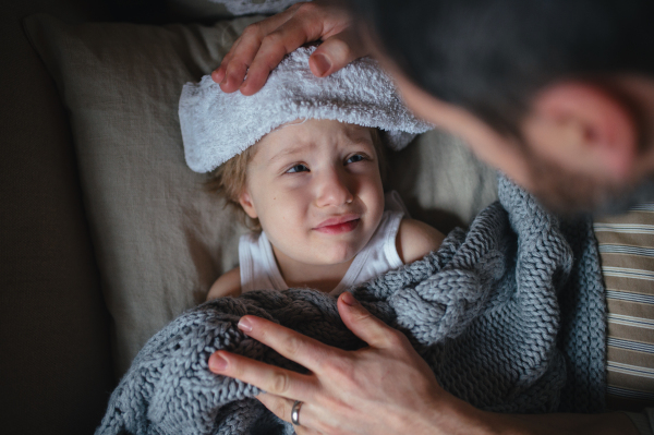 Father taking care of sick son with fever and cold, holdig cold cloth to his forehead. Boy lying on sofa, feeling tired.