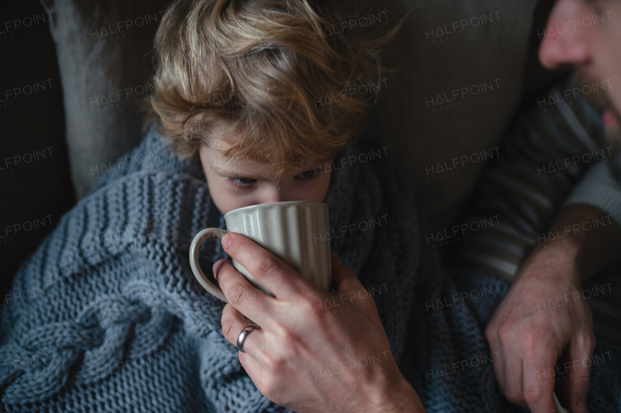 Father helping young son to drink warm tea from cup. Sick boy with cold lying under blanket and drinking herbal tea