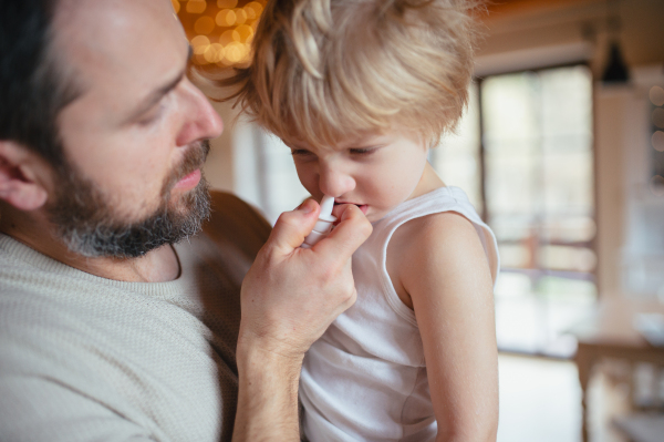 Sick boy with cold at home has stuffy nose. Father helping little son, using nasal spray in his nose