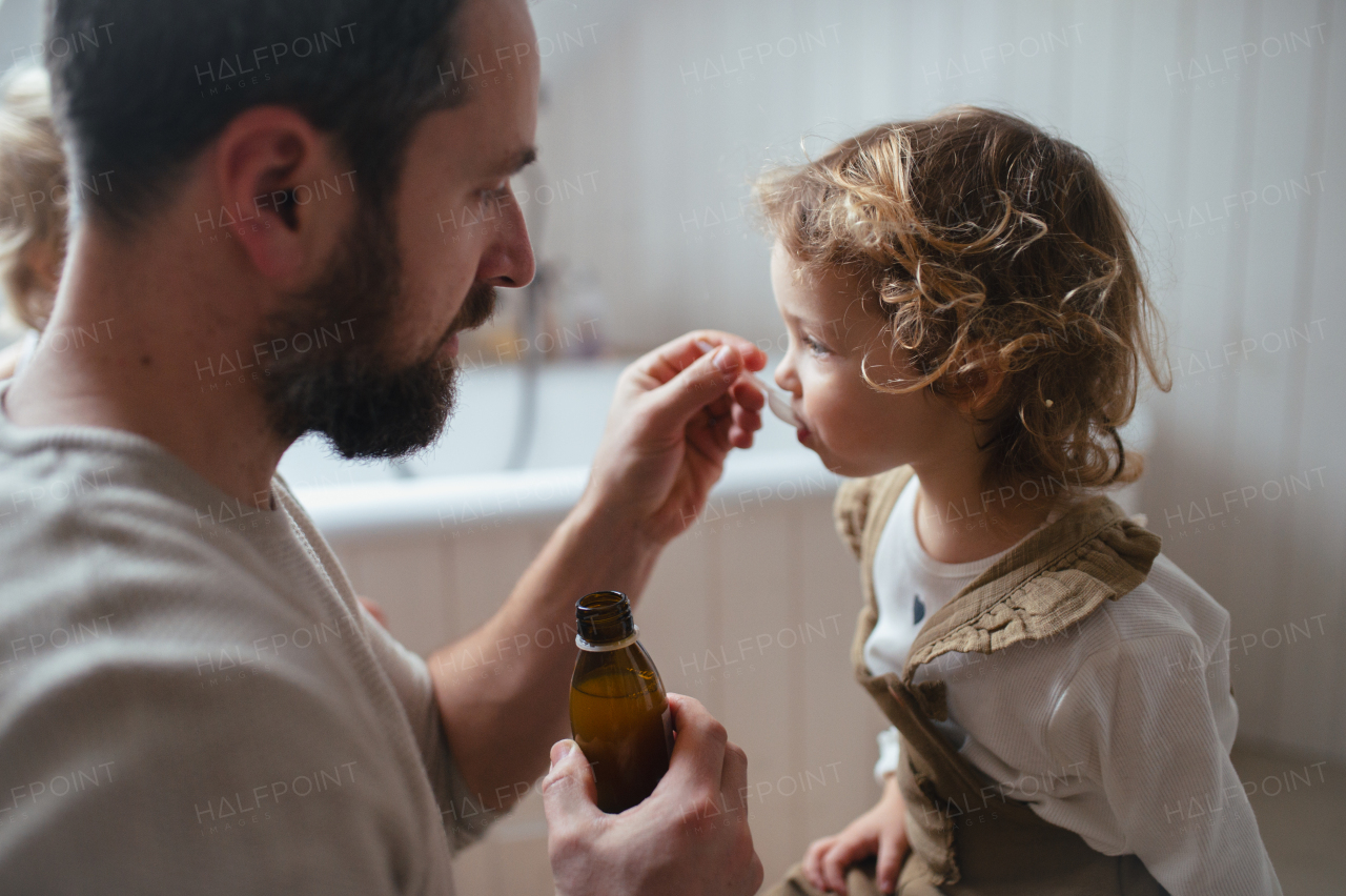 Father giving his daughter medicine on spoon, cough syrup or fever medicine. Sick girl with a fever and cold at home.