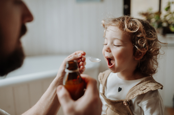 Father giving his daughter medicine on spoon, cough syrup or fever medicine. Sick girl with a fever and cold at home.