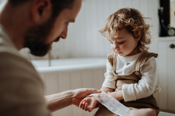 Father is bandaging his daughter's injured wrist in bathroom, concentrating while using a bandage