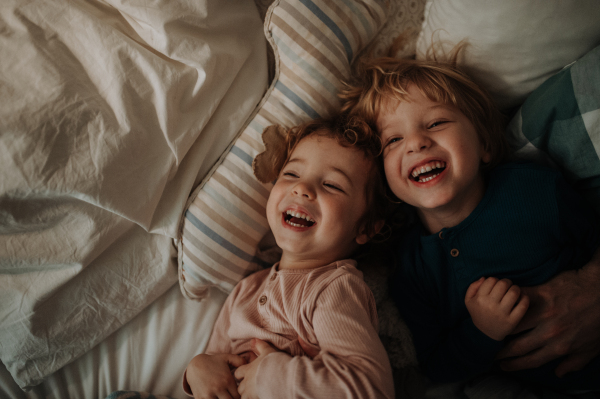 Candid portrait of siblings lying together in bed, looking at camera and laughing. Top view.