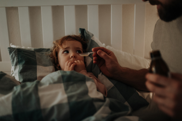 Father giving his daughter medicine on spoon, cough syrup or fever medicine. Sick girl with a fever and cold lying in bed.