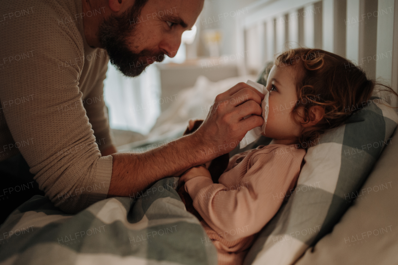 Sick girl with cold lying in the bed. Father helping little daughter to blow her nose