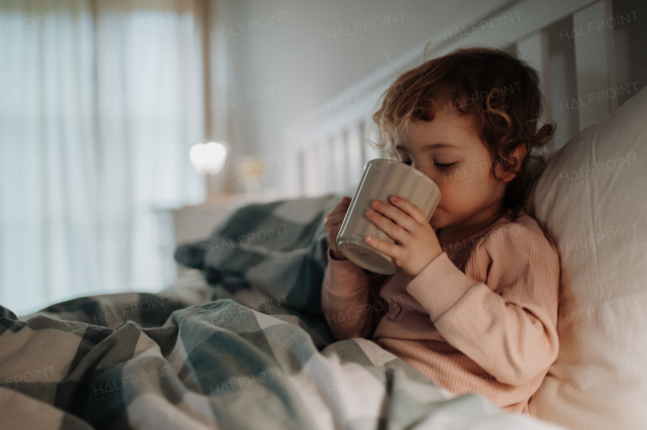 Little girl lying in the bed, coughing and holding tea cup. Sick girl with cold is drinking herbal tea