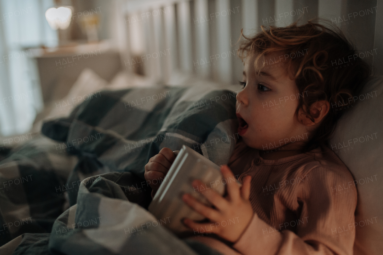 Little girl lying in the bed, coughing and holding tea cup. Sick girl with cold is drinking herbal tea