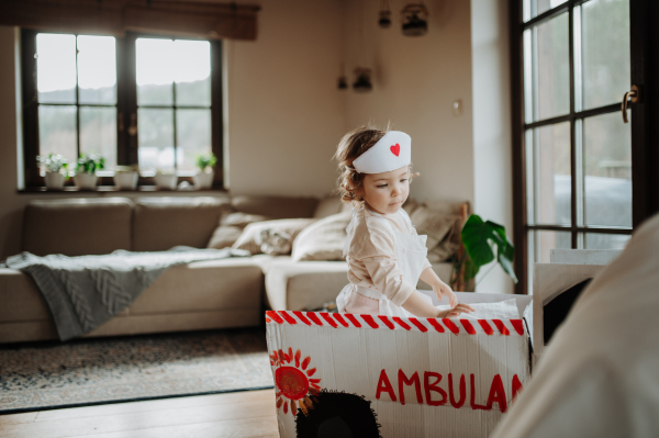 Girl in doctor uniform is playing hospital. Little girl as nurse sitting in cardboard ambulance