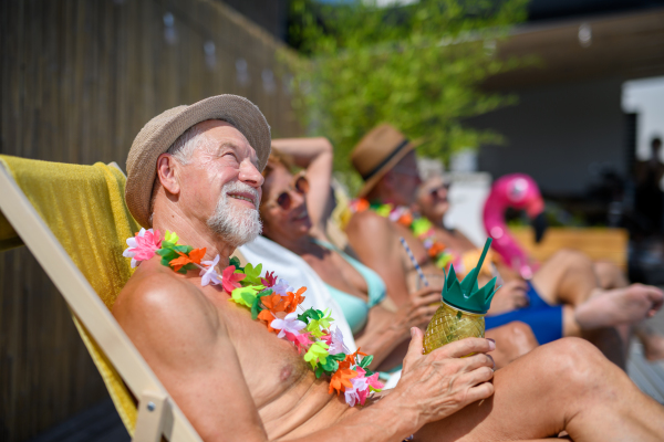 Group of cheerful seniors sitting lounge chairs by a swimming pool and sunbathing. Handsome senior man enjoying hot day outdoors in backyard with friends.