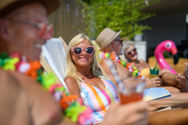 Group of cheerful seniors sitting lounge chairs by a swimming pool and sunbathing. Beautiful senior woman enjoying hot day outdoors in backyard with friends.