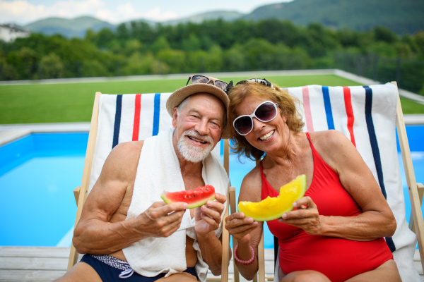 Beautiful elderly couple enjoying their vacation. Seniors having a romantic time, sitting by swimming pool in lounche chairs sunbathing.