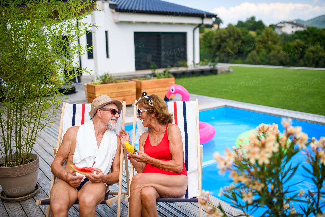 Beautiful elderly couple enjoying their vacation. Seniors having a romantic time, sitting by swimming pool in lounche chairs and eating watermelon.