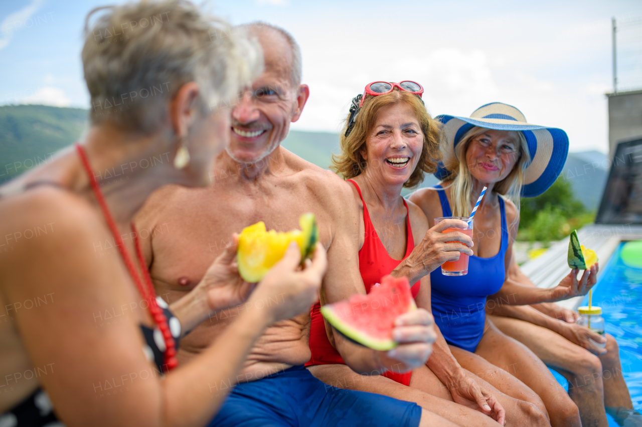 Group of cheerful seniors sitting on edge of pool with legs in water, eating watermelon, laughing. Elderly friends spending hot day by swimming pool, sunbathing.