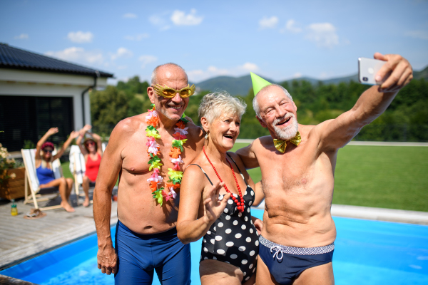 Group of cheerful seniors standing by pool taking selfie. Elderly friends spending a hot day by swimming pool, sunbathing.