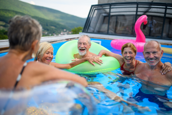 Group of cheerful seniors shaving fun in pool jumping, swiming and lounging on floats. Elderly friends spending a hot day by swimming pool.