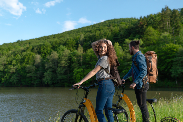 Travellers couple going on e-bike ride in nature. Tourist riding electric bicycles on easy trail road by lake. Young tourist spending romantic summer vacation oudoors.