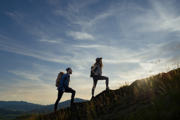 Travellers couple hiking on trail in nature with backpacks, during sunset. Young tourist spending romantic summer vacation oudoors. Side view, silhouette.