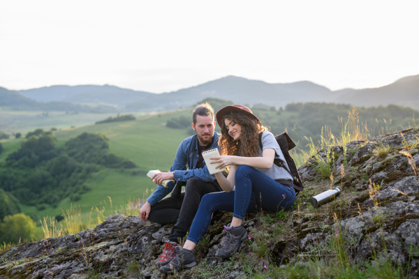 Travellers couple hiking on easy trail in nature with backpacks. Young tourist spending summer vacation oudoors, sitting on rocks, enjoying breathtaking view.