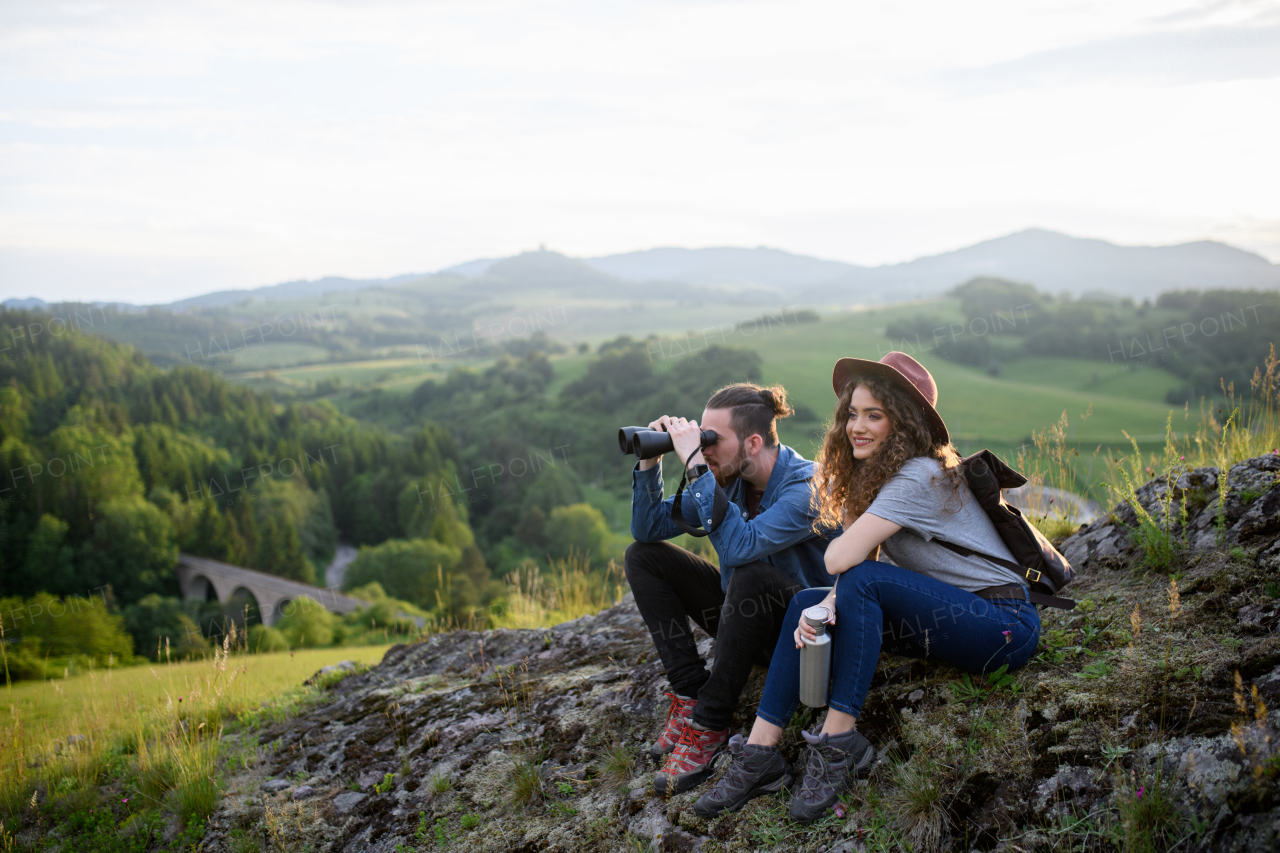 Travellers couple hiking on easy trail in nature with backpacks. Young tourist spending summer vacation oudoors, sitting on rocks, enjoying breathtaking view.