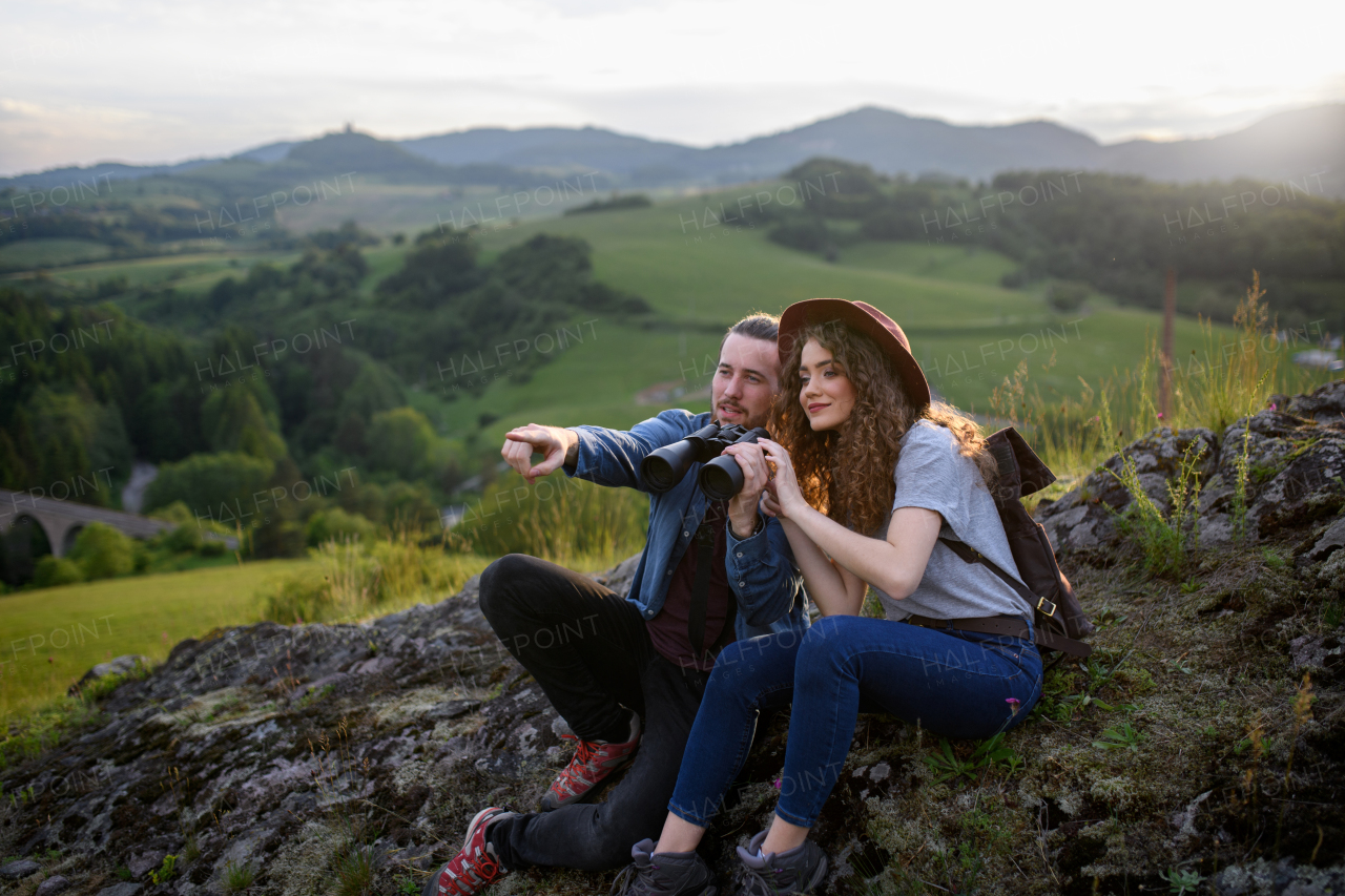 Travellers couple hiking on easy trail in nature with backpacks. Young tourist spending summer vacation oudoors, sitting on rocks, enjoying breathtaking view.