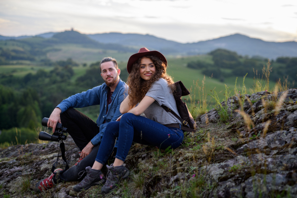 Travellers couple hiking on easy trail in nature with backpacks. Young tourist spending summer vacation oudoors, sitting on rocks, enjoying breathtaking view.