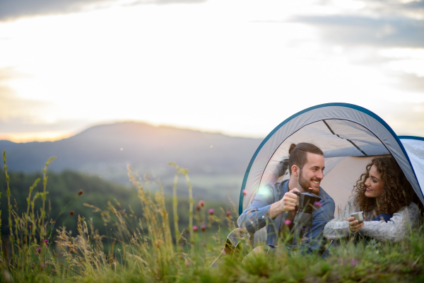 Travellers couple spending time outdoor, sleeping in tent shelter. Young tourist sitting in nature, resting, enjoying sunset. Romantic summer vacation in nature.