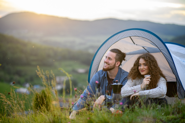 Travellers couple spending time outdoor, sleeping in tent shelter. Young tourist sitting in nature, resting, enjoying sunset. Romantic summer vacation in nature.