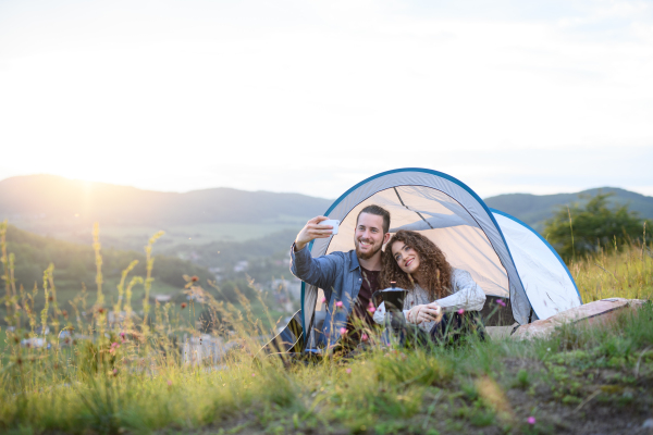 Travellers couple spending time outdoor, sleeping in tent shelter. Young tourist sitting in nature, resting, enjoying sunset. Romantic summer vacation in nature.