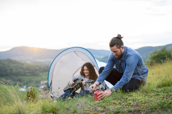 Travellers couple spending time outdoor, sleeping in tent shelter. Young tourist sitting in nature, resting, enjoying sunset. Romantic summer vacation in nature.