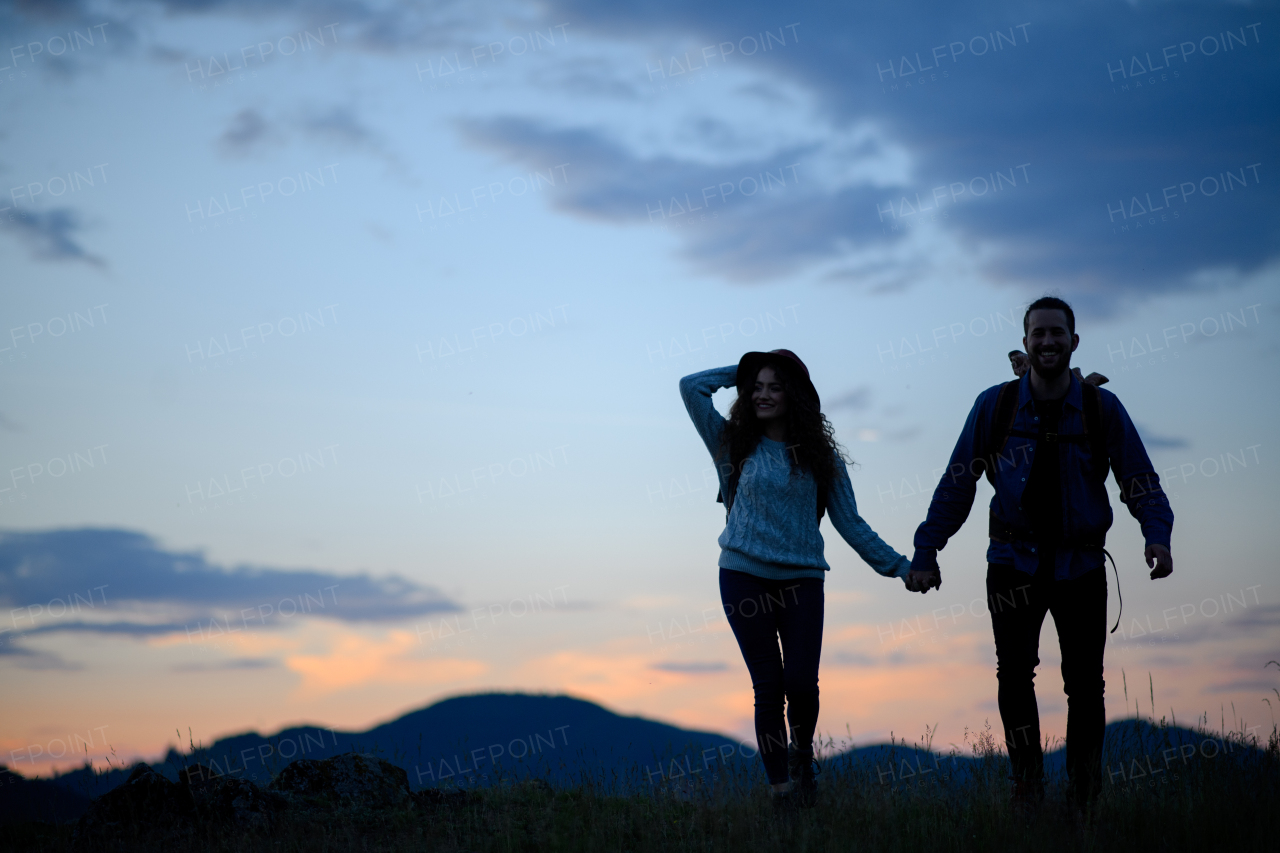 Travellers couple hiking on trail in nature with backpacks, during sunset. Young tourist spending romantic summer vacation oudoors. Side view, silhouette.