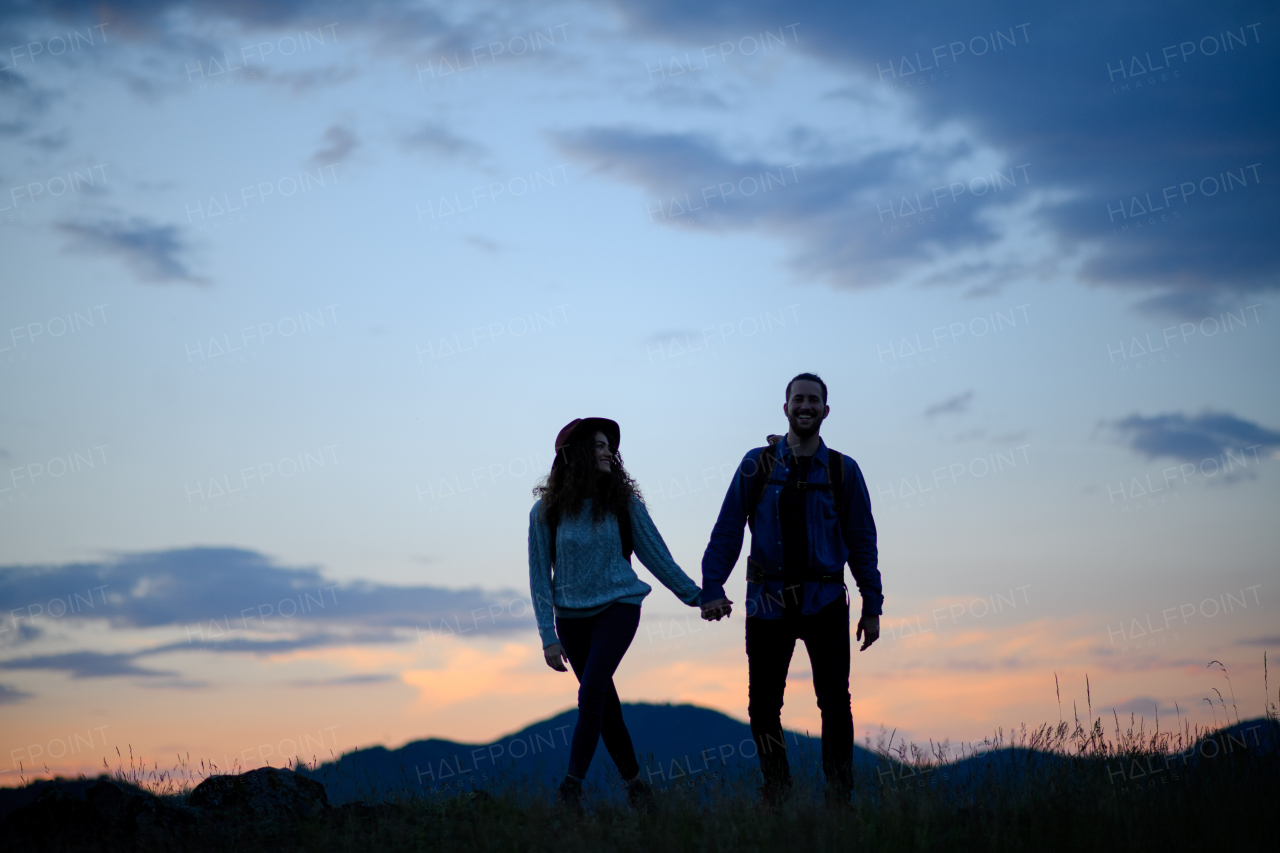 Travellers couple hiking on trail in nature with backpacks, during sunset. Young tourist spending romantic summer vacation oudoors. Holding hands.