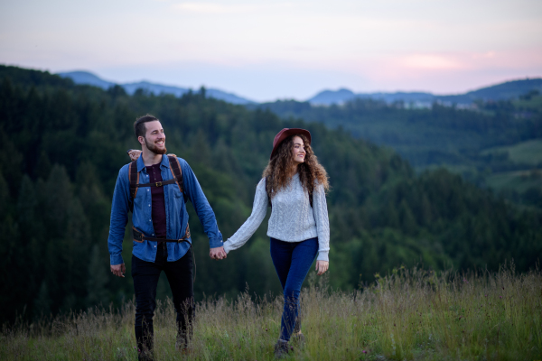 Travellers couple hiking on trail in nature with backpacks, during sunset. Young tourist spending romantic summer vacation oudoors. Holding hands.