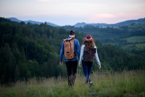 Travellers couple hiking on trail in nature with backpacks, during sunset. Young tourist spending romantic summer vacation oudoors. Holding hands.