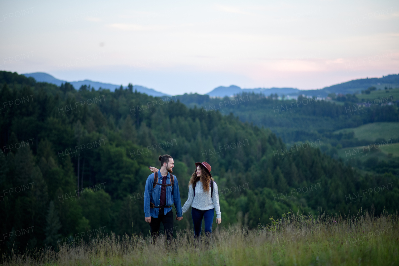 Travellers couple hiking on trail in nature with backpacks, during sunset. Young tourist spending romantic summer vacation oudoors. Holding hands.