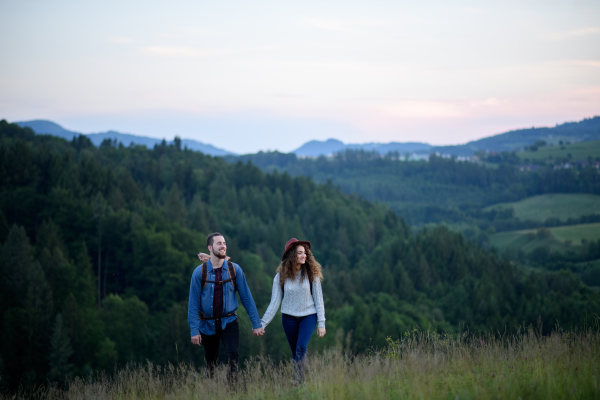 Travellers couple hiking on trail in nature with backpacks, during sunset. Young tourist spending romantic summer vacation oudoors. Holding hands.