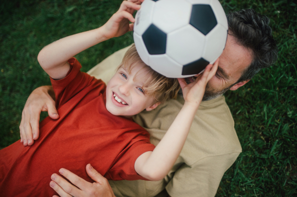 Dad lying on grass with son, laughing and having fun. Father and boy playing football, catching each other, falling to the ground. Fatherhood and Father's Day concept.